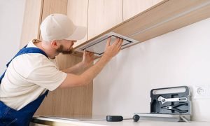 A professional handyman is focused on installing a range hood above the kitchen cabinetry in an apartment. Tools are neatly organized on the countertop, showcasing a tidy workspace.