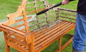 A gardener using a pressure washer to clean and restore a teak wood Lutyens style bench.